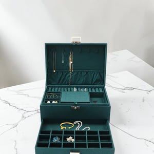 May include: A dark teal jewelry box with multiple compartments and a drawer. The box is open, revealing necklaces, bracelets, and rings. The interior is lined with a soft, dark teal fabric. The box has a gold-colored clasp and hinges.