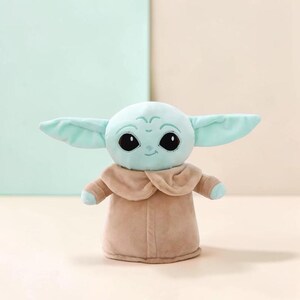 Puede incluir: Un peluche de un personaje con grandes ojos negros, orejas grandes y tez verde claro. El personaje lleva un abrigo marr&oacute;n. El juguete es un personaje popular de una franquicia de ciencia ficci&oacute;n.