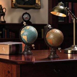 May include: Two decorative globe models with dark bases and gold accents sit on a wooden desk. One globe is teal and yellow, the other is bronze and orange. A vintage-style desk lamp with a brass finish is to the right.