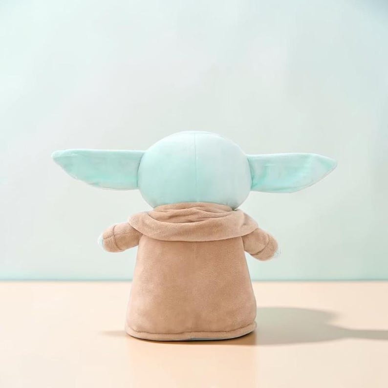 Puede incluir: Un peluche de un personaje con orejas grandes, cabeza verde claro y t&uacute;nica marr&oacute;n. El juguete se ve desde atr&aacute;s, mostrando su forma redondeada y textura suave. El juguete es un personaje popular de una franquicia de ciencia ficci&oacute;n.