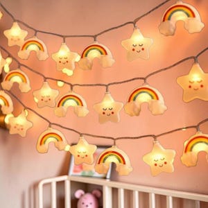 May include: String lights featuring smiling stars and rainbows with a warm, soft glow. The lights are strung on a brown wire and are suitable for a nursery or children's room. The lights are white with colorful rainbows.