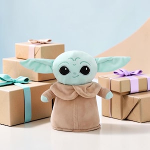 Puede incluir: Un peluche de un personaje con orejas grandes, ojos grandes y una t&uacute;nica marr&oacute;n. El juguete es de color verde claro y marr&oacute;n. Cajas de regalo con lazos en el fondo.