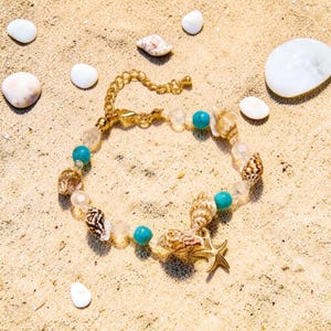 May include: A gold bracelet featuring a starfish charm, seashells, turquoise beads, and clear beads. The bracelet is displayed on sand, surrounded by seashells and white stones.