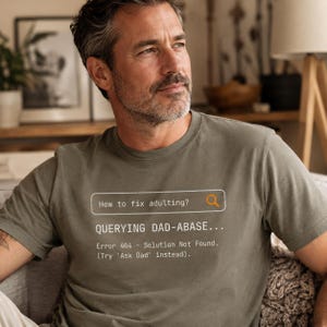 May include: Olive green t-shirt with the text "How to fix adulting?" and "QUERYING DAD-ABASE..." in a search bar design. The shirt also includes "Error 404 - Solution Not Found. (Try 'Ask Dad' instead)."