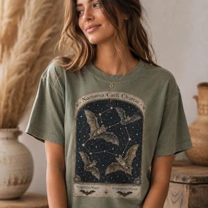 Può includere: T-shirt verde oliva con un'illustrazione vintage di pipistrelli e costellazioni. La grafica include il testo "Nocturna Caeli Chartae", "Vespertilio Major" e "Chiroptera Minor". Il design presenta un cielo notturno blu scuro con pipistrelli interconnessi.