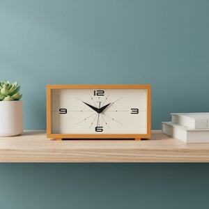 May include: A rectangular, orange-framed clock with a cream-colored face. Black numerals mark the 12, 3, 6, and 9 o'clock positions. The clock's hands are black, and the clock sits on a wooden shelf. A small succulent plant in a white pot is to the left.