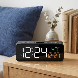 May include: A black digital alarm clock displaying the time as 12:24 PM, with the date and temperature shown in green and orange. The clock is on a wooden nightstand next to a blue pillow and decorative items.