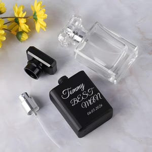Peut inclure: Un flacon de parfum en verre transparent avec un bouchon à facettes et un flacon de parfum rectangulaire noir mat portant le texte gravé blanc Tommy BEST man 06.05.2026, accompagné d'un bouchon noir séparé et d'un embout vaporisateur argenté.