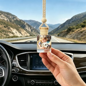 May include: Clear, square car air freshener with a photo of a child and a dog. The air freshener is suspended by a beige and gold braided cord, with a wooden bead and ring detail. The background shows a scenic mountain road.