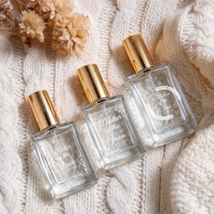 Peut inclure: Trois flacons de parfum rectangulaires en verre avec des bouchons dorés. Chaque flacon présente des inscriptions blanches gravées et des motifs floraux. On peut lire Mother of the Bride, Mother of the Groom 6.25.2026 et Mother of the Bride 6.25.2026.