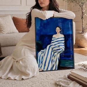 May include: A framed rectangular wall art print featuring a painting of a person with closed eyes, wrapped in a white and blue striped blanket, sitting on a dark blue sofa against a deep blue background, presented in a thin black frame.