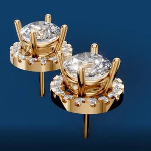 Puede incluir: Un par de pendientes de oro con grandes piedras centrales redondas y transparentes. Cada pendiente presenta un halo de piedras más pequeñas y brillantes y un poste para orejas perforadas. Fondo azul oscuro.