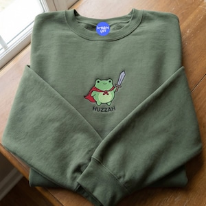 May include: An olive green sweatshirt with a crew neck and long sleeves. The front features an embroidered cartoon frog wearing a red cape and holding a sword, with the word "Huzzah" below. A blue sticker is on the upper back.