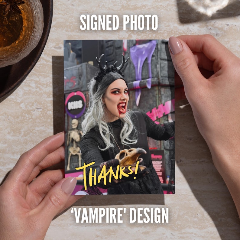 May include: A rectangular signed photo featuring a person with long silver hair, a black antler headband, and vampire-style makeup including red lips and fangs. The person wears a black top and holds a skull. The yellow text THANKS! is printed.