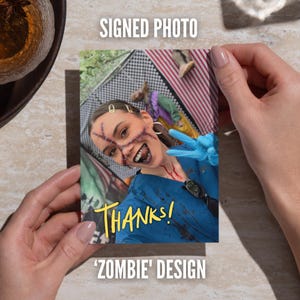 May include: A rectangular signed photo featuring a person in a blue medical scrub top with dark splatter marks, wearing blue gloves and makeup depicting facial scars and dark lips. The text THANKS! is printed in yellow on the lower portion.