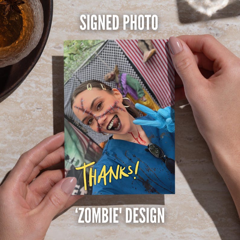 May include: A rectangular signed photo featuring a person in a blue medical scrub top with dark splatter marks, wearing blue gloves and makeup depicting facial scars and dark lips. The text THANKS! is printed in yellow on the lower portion.