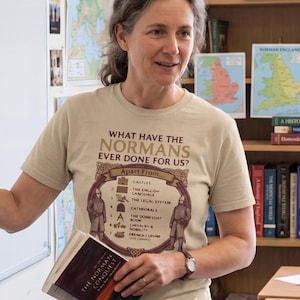 May include: A beige short-sleeved t-shirt featuring a graphic print with the text "WHAT HAVE THE NORMANS EVER DONE FOR US?" and a list of seven historical contributions including castles, the English language, and cathedrals, flanked by two figures.