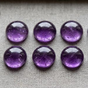 Op de afbeelding: Acht ronde, gepolijste amethist cabochons gerangschikt in twee rijen van vier. Elke paarse edelsteen heeft een glad, koepelvormig oppervlak met zichtbare natuurlijke insluitingen. Een deel van een metalen liniaal is zichtbaar.