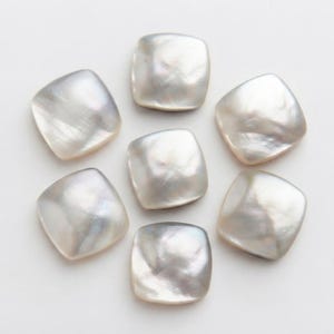 Può includere: Sette bottoni quadrati con angoli arrotondati, ognuno caratterizzato da una superficie liscia e iridescente in madreperla color bianco sporco. I bottoni sono disposti in un motivo circolare, mostrando variazioni naturali nella luce.