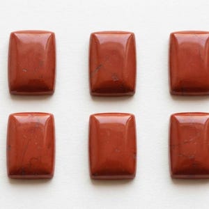 Op de afbeelding: Zes rechthoekige rode jaspis cabochons gerangschikt in twee rijen van drie. Elke steen heeft een glad, gepolijst oppervlak met een licht gewelfde bovenkant en subtiele, donkere natuurlijke aders zichtbaar in het dieproodbruine materiaal.