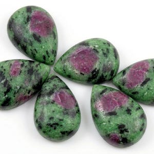 Può includere: Cinque gemme di rubino in zoisite lucidate a forma di goccia disposte in gruppo. Ogni pietra presenta una superficie liscia e lucida con un motivo maculato di zoisite verde vibrante, inclusioni di rubino magenta intenso e piccole macchie nere.