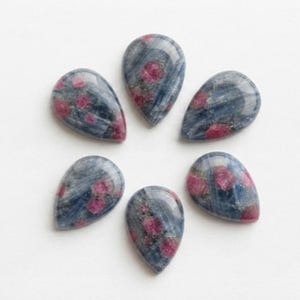 May include: Six teardrop-shaped cabochons arranged in a circular pattern. Each stone features a polished, smooth surface with a deep blue, fibrous base colour and distinct, scattered inclusions of vibrant, opaque pinkish-red mineral spots.