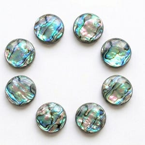 May include: Seven circular buttons made of iridescent abalone shell, arranged in a circular pattern. Each button features a unique, swirling pattern of shimmering blue, green, and pink hues with a polished, smooth surface and a rounded edge.