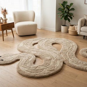 May include: A large, beige, snake-shaped area rug with a textured, tufted surface. The rug features a winding, serpentine body with a coiled tail and a distinct head with dark, embroidered eyes and a forked tongue, laid flat on a wooden floor.
