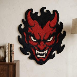 May include: A tufted wall hanging featuring a red and black demon face with horns and sharp teeth. The design is surrounded by a black flame-like border, creating a bold, textured appearance. The piece is crafted with a dense, looped pile texture.