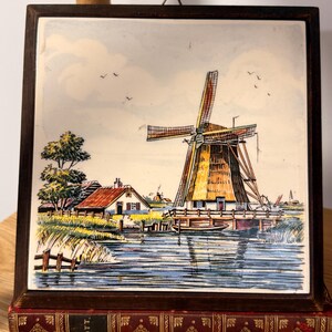 May include: A framed ceramic tile with a Dutch windmill scene. The artwork shows a windmill with a thatched roof, a small house, and water. The dark brown frame surrounds the tile, which rests on a decorative book.