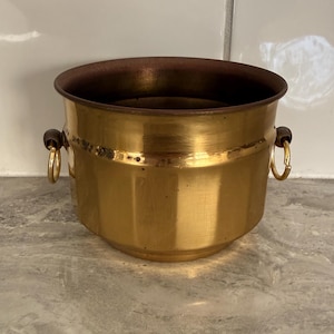 May include: A polished brass bucket with a wide, open top and a dark interior. The container features two side handles, each consisting of a small dark wooden bead attached to a circular brass ring, providing a functional grip for the vessel.