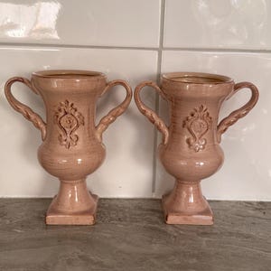 May include: A pair of matching dusty pink ceramic urns featuring two curved handles on each side, a square base, and a raised decorative emblem on the front. The surface has a subtle, textured finish with darker pink highlights throughout.