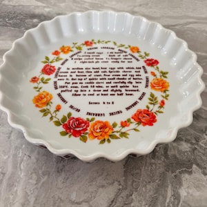 May include: A white ceramic fluted quiche dish featuring a central recipe for Quiche Lorraine printed in black text, surrounded by a decorative wreath of orange, yellow, and red roses. The text includes ingredients and baking instructions.