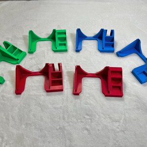 May include: An assortment of colorful, 3D-printed plastic objects in green, red, and blue. The objects have a bridge-like design with a shelf-like structure on one side. They are arranged on a white, textured surface.