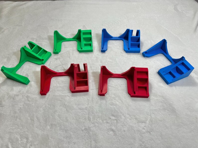 May include: An assortment of colorful, 3D-printed plastic objects in green, red, and blue. The objects have a bridge-like design with a shelf-like structure on one side. They are arranged on a white, textured surface.