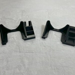 May include: Two black plastic brackets, each with an arched support and a rectangular base with three shelves. The brackets are designed to hold an object, likely for display or support. The image is taken from a slightly elevated angle.