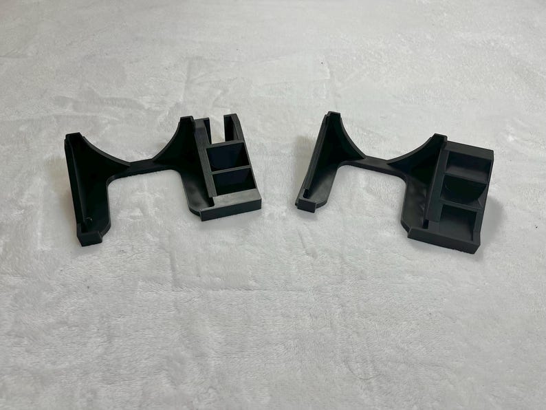 May include: Two black plastic brackets, each with an arched support and a rectangular base with three shelves. The brackets are designed to hold an object, likely for display or support. The image is taken from a slightly elevated angle.