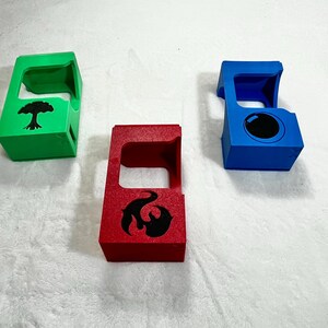 May include: Three colorful, geometric card holders. One is green with a black tree design, another is red with a black flame design, and the third is blue with a black circle. The holders are made of a solid material.