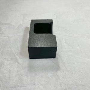 May include: A black, textured box with a rectangular opening on top. The box has a small, angled side and a dark interior. The box is sitting on a white, textured surface.