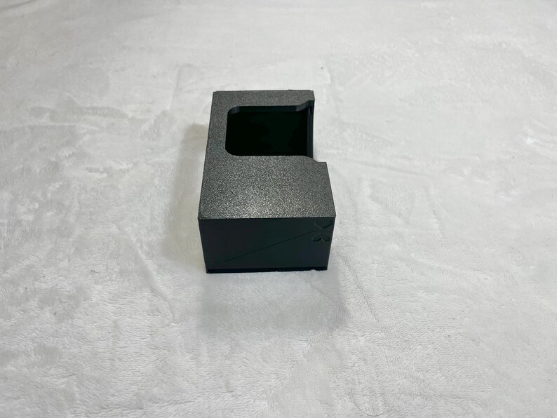 May include: A black, textured box with a rectangular opening on top. The box has a small, angled side and a dark interior. The box is sitting on a white, textured surface.