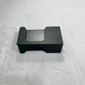 May include: A small, rectangular, black card holder with a textured surface. The holder has a cutout on one side and a slightly raised edge on the other. The item is sitting on a white, fluffy surface.