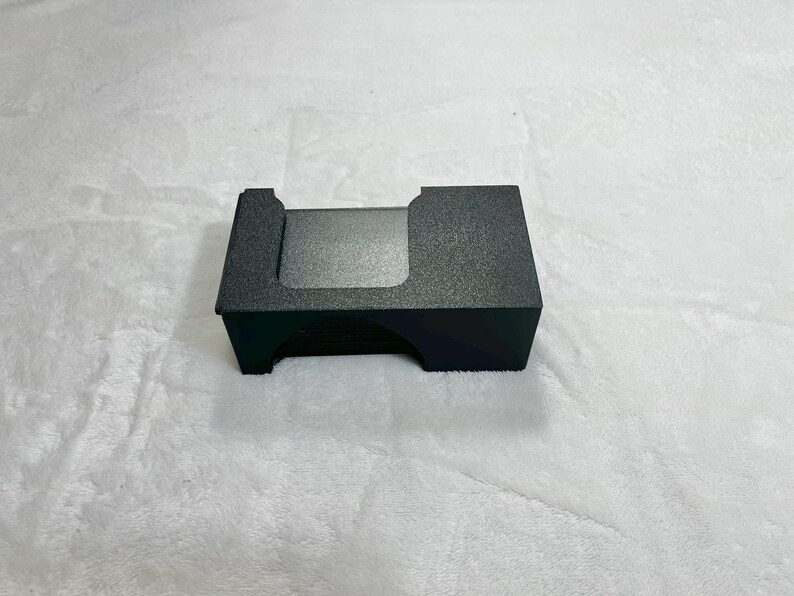 May include: A small, rectangular, black card holder with a textured surface. The holder has a cutout on one side and a slightly raised edge on the other. The item is sitting on a white, fluffy surface.