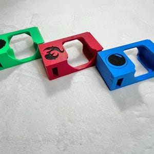 May include: Three colorful, rectangular objects with cut-out centers. One is green with a black tree design, another is red with a black emblem, and the third is blue with a black circle. The objects are likely cases or holders.