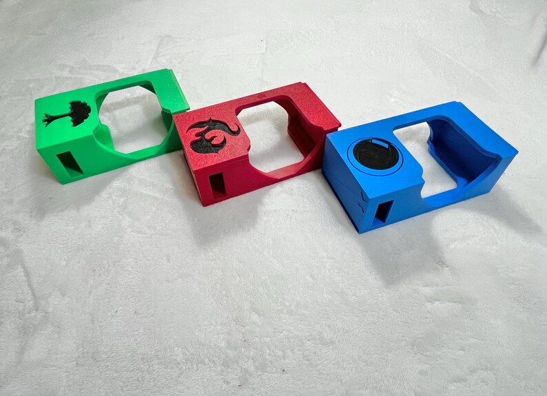 May include: Three colorful, rectangular objects with cut-out centers. One is green with a black tree design, another is red with a black emblem, and the third is blue with a black circle. The objects are likely cases or holders.