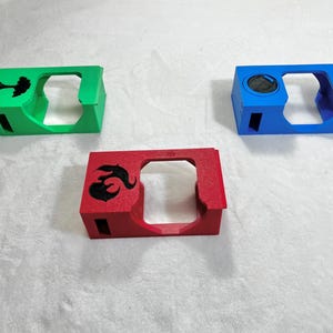 May include: Three colorful, rectangular cases with cut-out sections. One is green with a tree silhouette, another is red with a black flame design, and the third is blue with a circular detail.