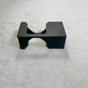 May include: A black, textured plastic stand with a rectangular top and a curved archway underneath. The stand is designed to hold an item, with a cutout on one side. The object is on a white surface.