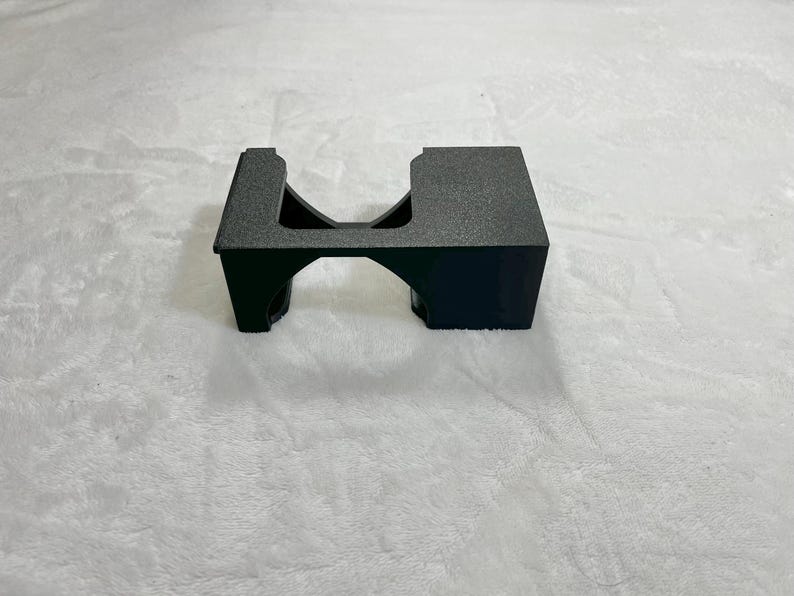 May include: A black, textured plastic stand with a rectangular top and a curved archway underneath. The stand is designed to hold an item, with a cutout on one side. The object is on a white surface.
