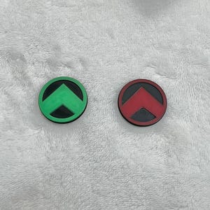 May include: Two circular, rubber-like objects. One is green with a black arrow, the other is red with a black arrow. Both have a black border. The objects are on a white, textured surface.