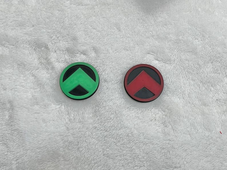 May include: Two circular, rubber-like objects. One is green with a black arrow, the other is red with a black arrow. Both have a black border. The objects are on a white, textured surface.