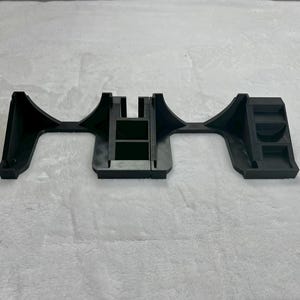 May include: A black plastic bracket with a unique shape, featuring a central rectangular section and curved, flared ends. The bracket has a series of rectangular cutouts and raised sections, suggesting a structural or mounting component.